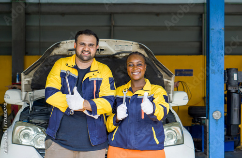 Two mechanic in uniform and protective glove raise finger thumbs up, stand in front of a car with an open hood, smile while holding wrench tool.