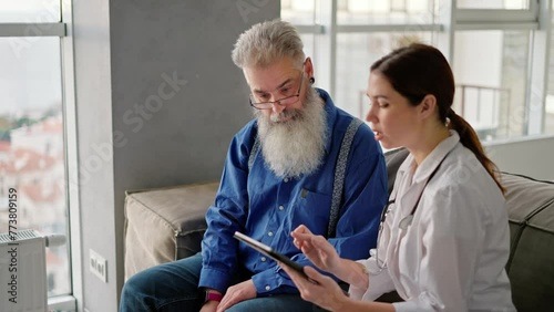 A brunette woman in a white medical coat shows a man with gray hair with glasses and a lush beard on a tablet their names and recipes for drugs to improve health after a home examination with a doctor photo
