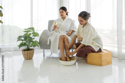 Happy young asian woman in bathrobe with glass of champagne having fun with foot bath from cosmetologist during procedure in spa salon.