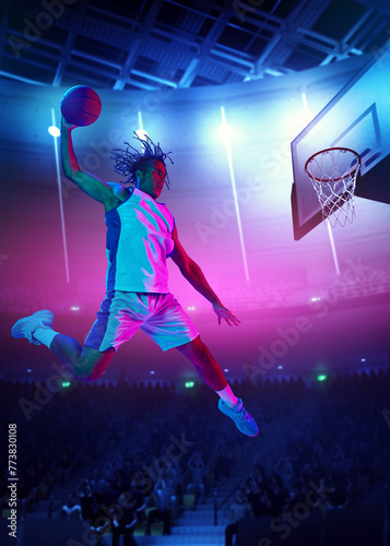 Slam dunk. Competitive young man, basketball player throwing ball into basketball hoop in a jump on gradient stadium background with spotlights. Concept of sport, action, game, competition, tournament © master1305