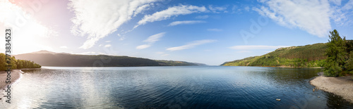 Panoramic drone shot of Loch Ness, where the gentle rays of the sun bathe the tranquil waters and pebbled shores in a warm glow, with dense forests and hills framing the scene in natural splendor © Artem