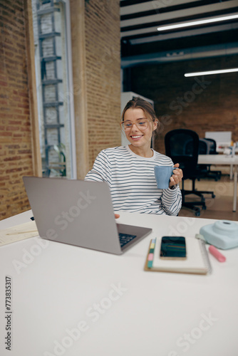 Young business woman working on laptop and drink coffee while sitting the desk in stylish office 