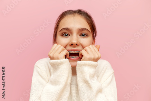 scared teenage girl with braces worries and bites nails on pink isolated background, excited child nervous in stress