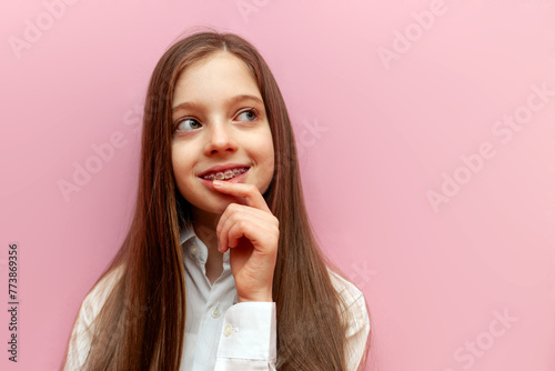 pensive teenage girl in a white shirt and braces thinks and plans on a pink isolated background, puzzled child dreams and imagines