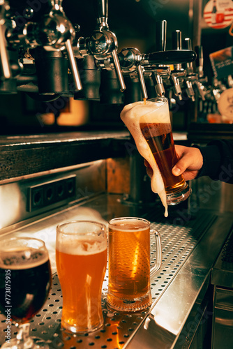 Photo of bartender pouring light, foamy beer from tap into pint glass at bar with multiple beer dispensers. Serving craft beer from tap. Concept of alcohol drinks, nightlife, festivals, Oktoberfest.