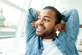 happy relaxed african american man with braces resting and looking out the window in a white room, the man smiles and dreams while looking away with his hands behind his head