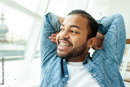 happy relaxed african american man with braces resting and looking out the window in a white room, the man smiles and dreams while looking away with his hands behind his head