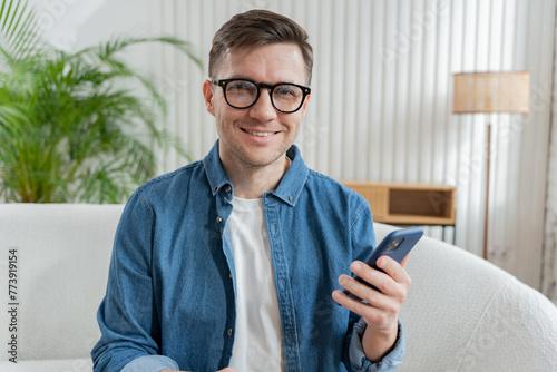A cheerful man holding a smartphone radiates satisfaction, perched comfortably in an airy, plant-filled living area.