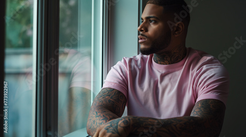 the stillness of the room, a man in a pink t-shirt leans against a window frame, his arms a canvas of intricate tattoos that reflect his innermost thoughts and experiences