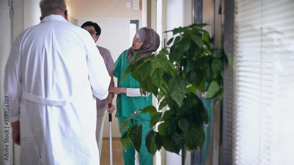 Female caregiver helping her female patient with a leg injury, holding her hand, supporting her. Young Muslim nurse wearing hijab in a uniform supports a mature elderly woman while she walks with Female caregiver helping her female patient with a leg injury, holding her hand, supporting her. Young Muslim nurse wearing hijab in a uniform supports a mature elderly woman while she walks with