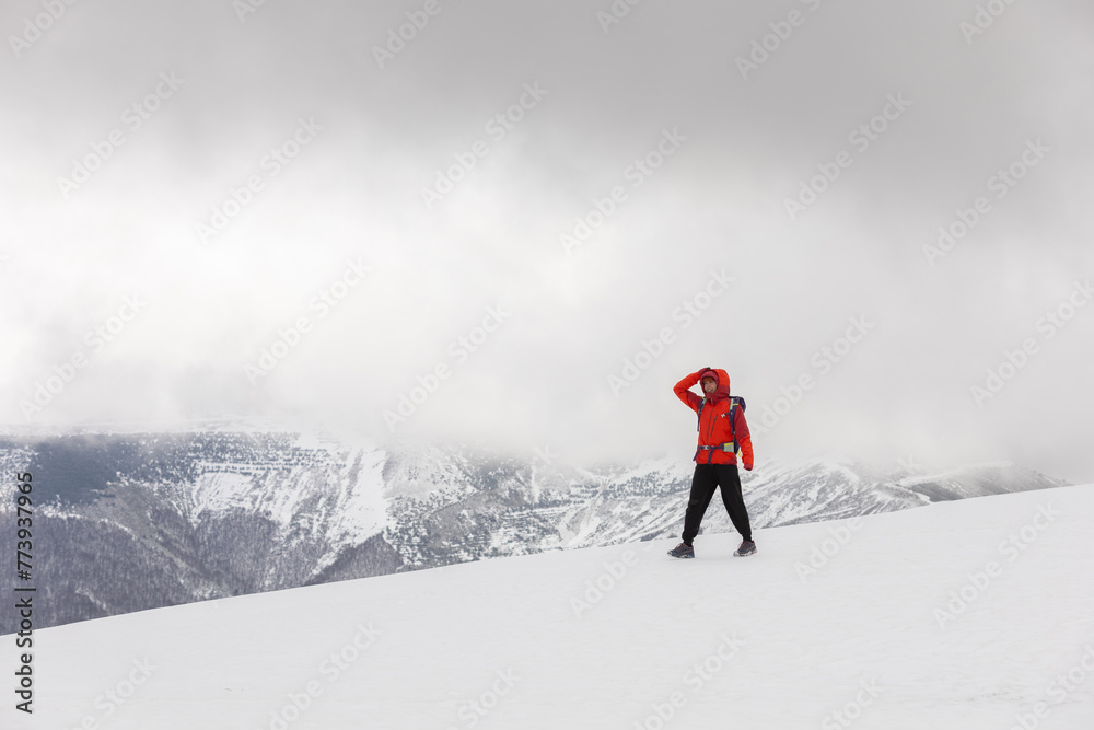 A man in an orange jacket stands on a snowy mountain peak, looking out at the landscape. The scene is serene and peaceful, with the man's gaze focused on the horizon
