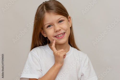 Portrait of caucasian little girl with open wide smile holding finger on cheek looking at camera