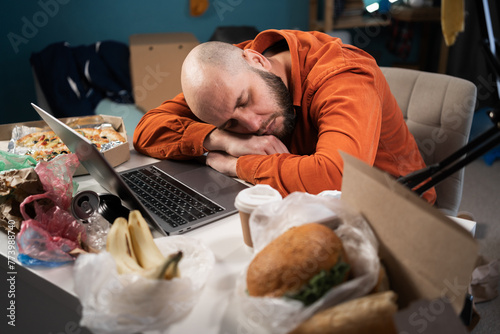 Exhausted millennial man sleeping on desk near laptop, working in messy room, tired of overworking. Workaholic suffering from chronic fatigue at workplace