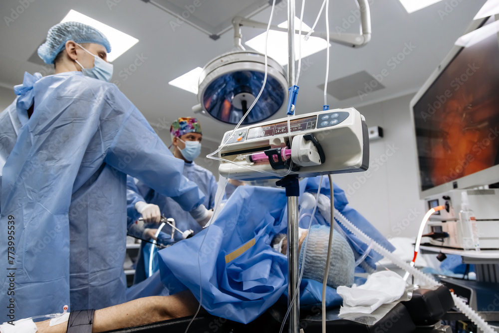 Experienced surgeons in medical gowns, caps, masks and gloves perform an operation in a modern clinic. Group of professional doctors using modern medical equipment in operating room. Experienced surgeons in medical gowns, caps, masks and gloves perform an operation in a modern clinic. Group of professional doctors using modern medical equipment in operating room.
