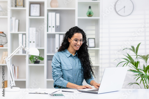 Smiling Hispanic woman focused on work at her home office, exuding professionalism and confidence in a bright setting. © Liubomir