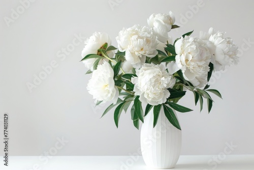 White peonies in white vase on table. White vase is overflowing with an abundance of pristine white flowers. The delicate petals and green stems create a beautiful arrangement inside the vase.