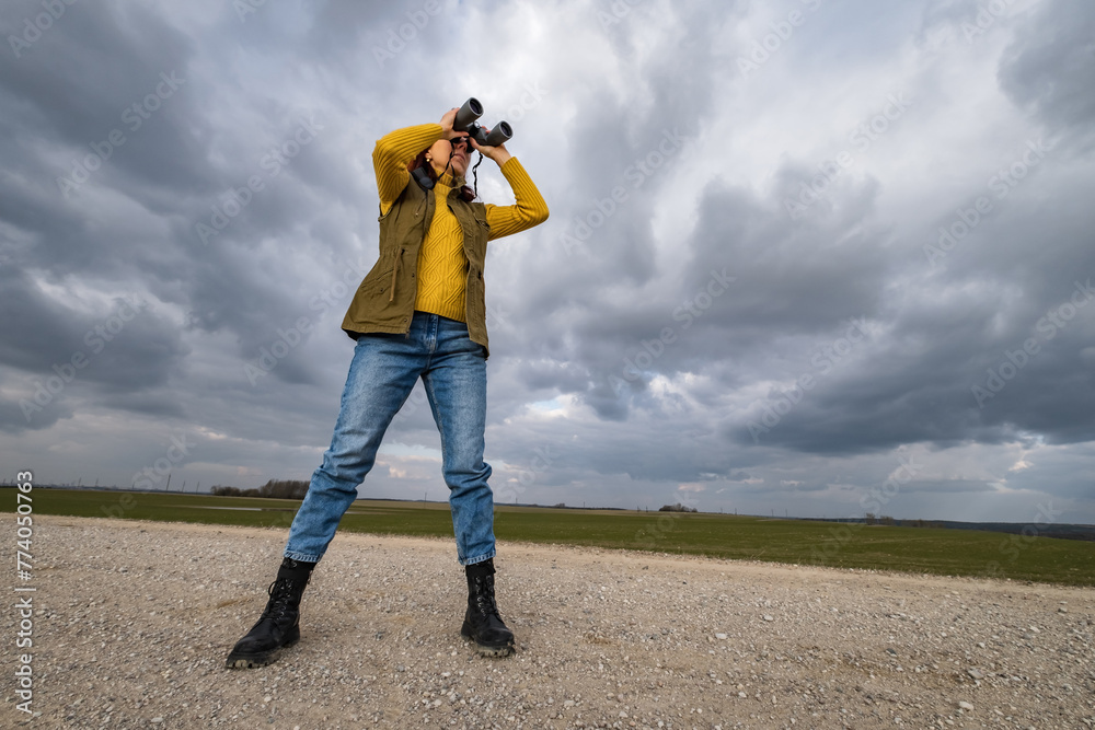 female ornithologist birdman watches birds with binoculars on dirt gravel road against a background of a stormy sky female ornithologist birdman watches birds with binoculars on dirt gravel road against a background of a stormy sky