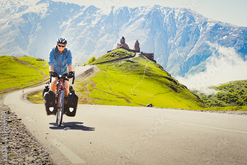 Male cyclist on touring bicycle adventure cycle visits Gergeti trinity church with mountains background. Cycling holidays and travel around caucasus mountain range