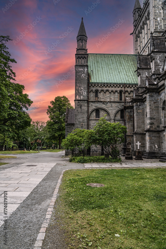 Sunset at Nidaros Cathedral courtyard in Trondheim, Norway. The evening sky ablaze with sunset hues behind the Cathedral spire, with gravestones scattered across the lush garden.