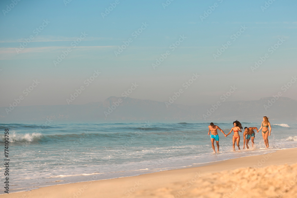 Couple With Friends In Swimwear On Vacation Holding Hands Running Along Beach Shoreline Couple With Friends In Swimwear On Vacation Holding Hands Running Along Beach Shoreline