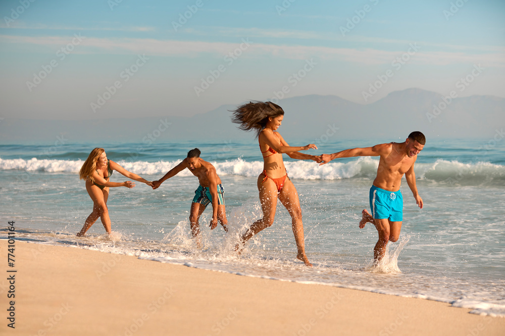 Couple With Friends In Swimwear On Vacation Holding Hands Running Along Beach Shoreline 