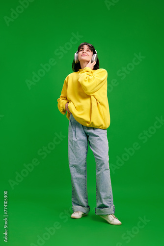 Full-length portrait of overjoyed woman in oversize outfit listening music in headphones against vibrant green studio background. Concept of human emotions, fashion, beauty, style, recreation, hobby. © Lustre Art Group 