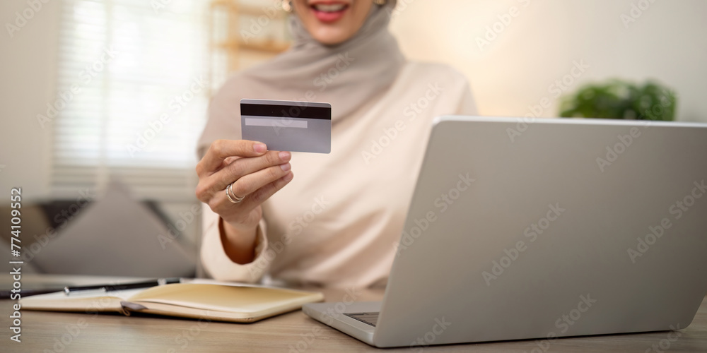 Young happy muslim woman in hijab at home using laptop shopping online with credit card while sitting on desk Young happy muslim woman in hijab at home using laptop shopping online with credit card while sitting on desk
