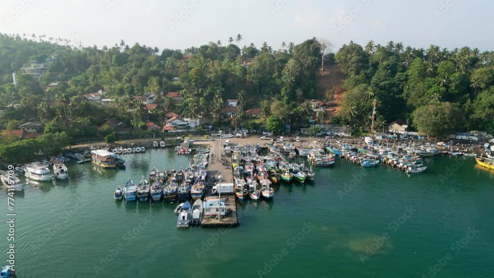 Aerial drone of Mirissa harbor with bustling activity around colorful fishing, motor boats moored in calm blue Indian Ocean water, green tropical trees surround busy fisherman port in Asia. Aerial drone of Mirissa harbor with bustling activity around colorful fishing, motor boats moored in calm blue Indian Ocean water, green tropical trees surround busy fisherman port in Asia.