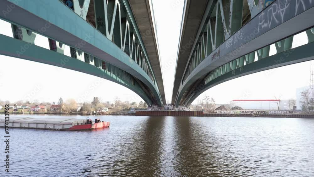 A cargo barge passes beneath a steel bridge, framed by symmetrical bridge structures over a calm river A cargo barge passes beneath a steel bridge, framed by symmetrical bridge structures over a calm river