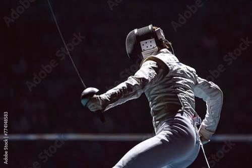 A man in a white fencing suit and a black fencing mask stands with a sword while sparring on the platform.