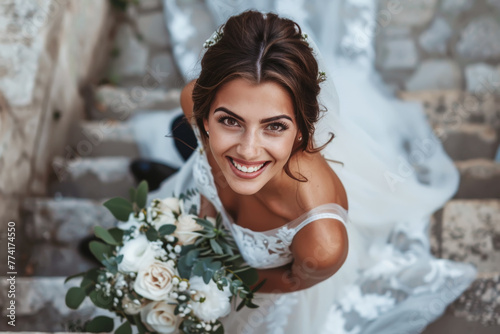 Hermosa novia con vestido blanco con encaje y pedreria, llevando un ramo de flores en tonos claros, mirando hacia arriba en unas escaleras antiguas de subida a la iglesia photo