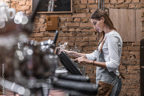 Young caucasian woman in apron working with cash box in coffee shop behind the counter. Female worker, barista, waitress, manager, administrator checking bill order on digital cash register. photo