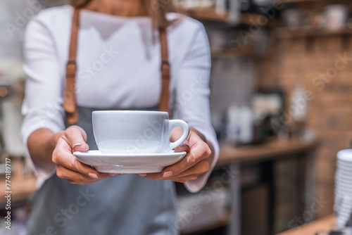 Young woman barista holding coffee cup serving client at coffee shop. Student working part-time in cafe, made order, gives mug for customer, wearing uniform apron. Cropped shot of white cup mug photo