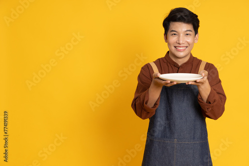 Young food waiter Asian man holding empty white plate or dish isolated on yellow background
