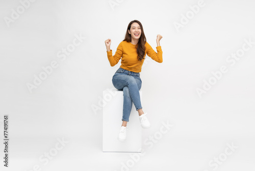Attractive beautiful Asian female sitting on white wood box and hands up raised arms from happiness, Excited woman winner success concept