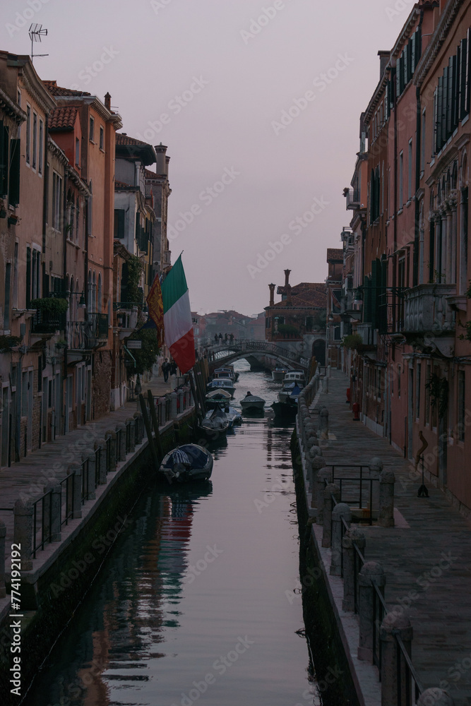 Typical narrow canal surrounded by buildings with boats during evening twilight, Venice, Veneto, Italy