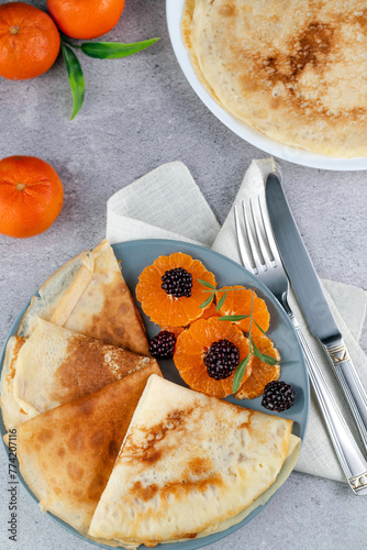 On a gray table there are pancakes with fruit, a fork and a knife, tangerines.