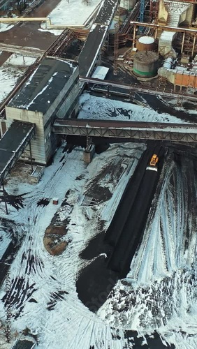 Yellow excavator smoothes black ash from coal under the bridge in the territory of the heating power station. Aerial view. Vertical video photo