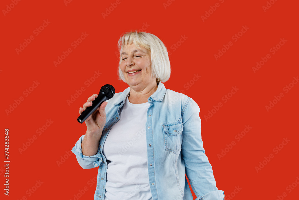 Old pretty happy woman singing a song with a microphone, isolated on red background, studio portrait. Music lover enjoying music Old pretty happy woman singing a song with a microphone, isolated on red background, studio portrait. Music lover enjoying music