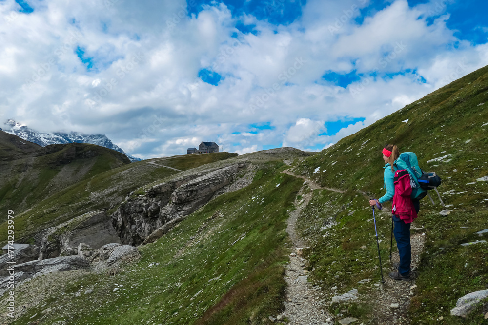 Hiker woman with panoramic view of alpine cottage Hagener Huette surrounded by mountain peaks in High Tauern National Park, Carinthia, Austria. Idyllic hiking trail along high altitude alpine meadow