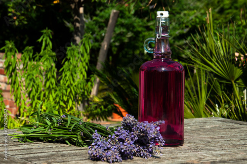 Homemade lavender syrup in a buckled bottle, placed on an old wooden table outdoors in bright summer sunshine. A bouquet of lavender flowers accompanies the syrup, adding to the charming, rustic feel.