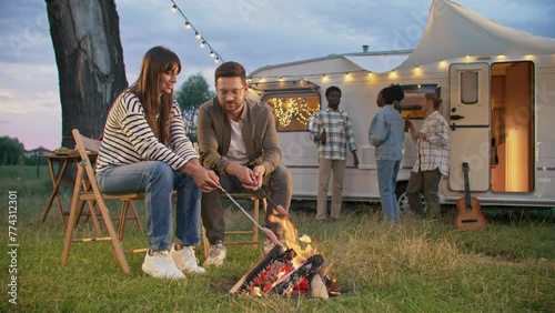 Happy couple sitting near campfire and roasting sausages during conversation. Group of friends having picnic outdoors. People drinking beers and dancing by music before white trailer in background.