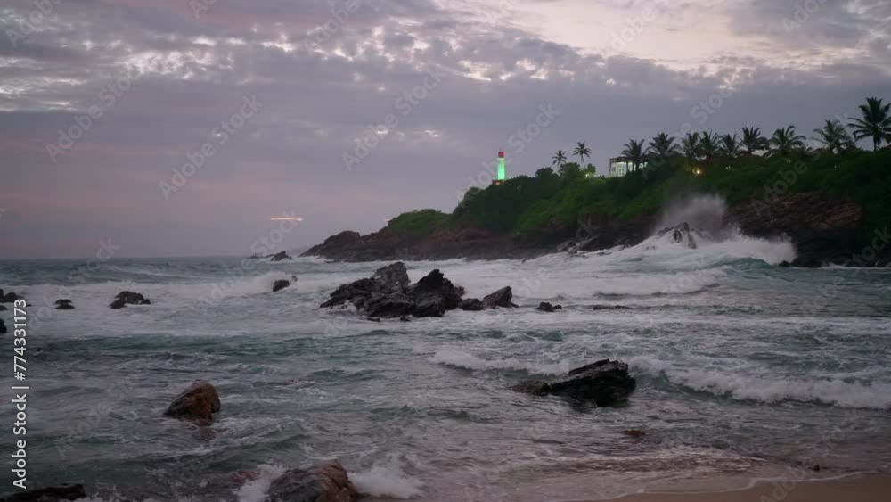 Twilight at rocky sea coast with waves crashing. Waves break on stones at dusk. Lighthouse beams green in tropical landscape. Purple sky over ocean after sunset. Nature scene with water motion. Slowmo Twilight at rocky sea coast with waves crashing. Waves break on stones at dusk. Lighthouse beams green in tropical landscape. Purple sky over ocean after sunset. Nature scene with water motion. Slowmo