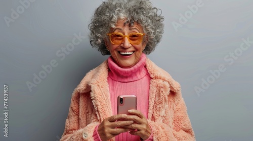 Joyful senior lady with curly gray hair and orange glasses holding a phone. © AdriFerrer