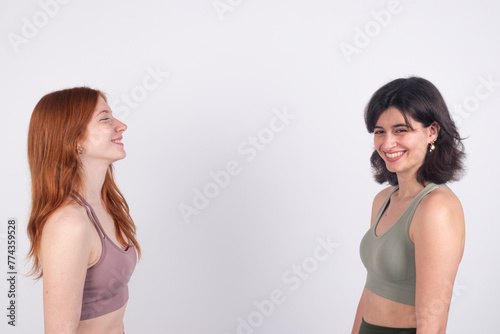 Two young women smiling and facing each other against a white background, expressing happiness and friendship.