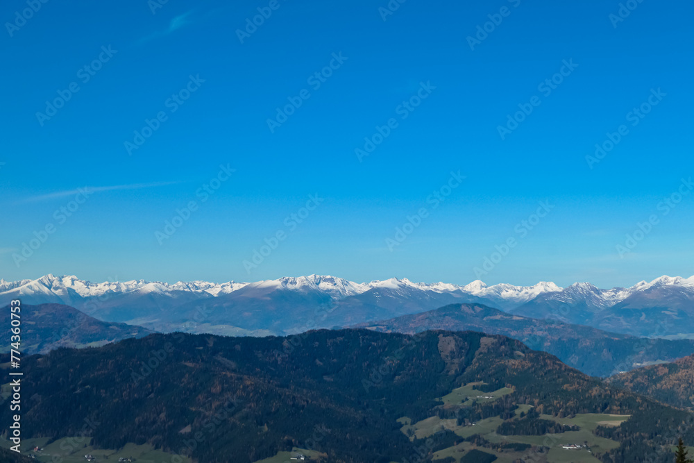 Panoramic view of snow capped mountain ridges of Woelzer Tauern seen from Grebenzen, Gurktal Alps, Styria, Austria. Calm serene atmosphere in Austrian Alps. Idyllic forest in foreground. Wanderlust