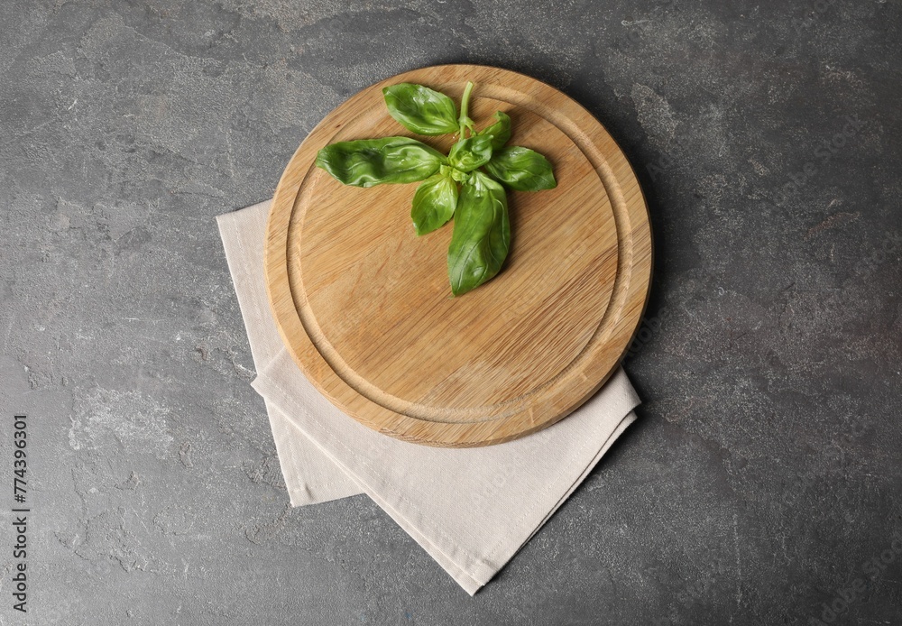 Wooden cutting board, fresh basil and napkin on grey table, top view