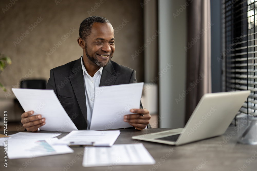 African American businessman in a suit smiles while reviewing papers in an office setting with a laptop and other documents on his desk. African American businessman in a suit smiles while reviewing papers in an office setting with a laptop and other documents on his desk.