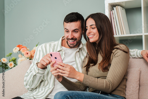 Fotografi Caucasian couple having fun using a mobile phone to browse on internet at living room sofa