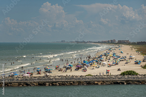 Port Canaveral, Florida, USA - July 30, 2023: Long sandy beach, full of bathers in water and on sand, stretches out as far as one can see, south  of Fisherman Pier under blue cloudscape